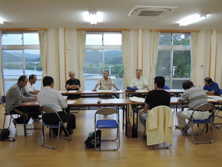 語る中平副会長。聞く参加者。 2017年第3回定例会の様子。中央に熱弁する中平副会長。ロの字に机が並んでいて、11人が座って話を聞いています。中平さんは両手を机にひろげてついて、座って話しています。その後には背の高い窓が3つ並んでいて、台風が接近している空模様がよく分かります。四万十川の水が増えているのも見えます。